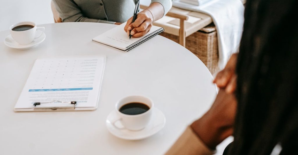 From above crop multiracial female interviewer or psychologist in formal wear asking questions and taking notes in planner while talking to black man and sitting together at table with coffee